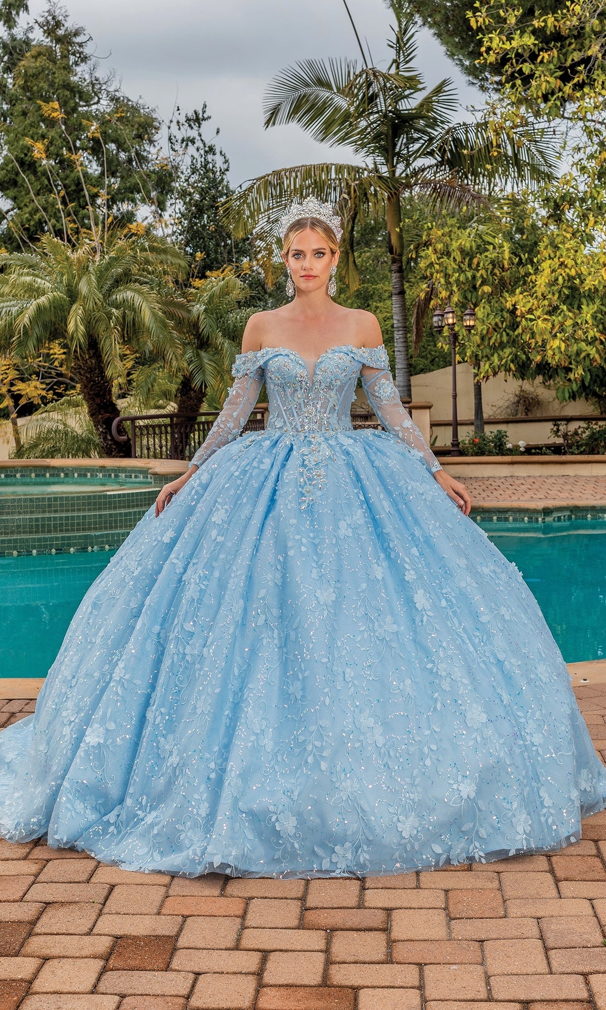 A woman stands outdoors by a pool wearing the Quinceanera Dress 1890 By Dancing Queen, a light blue off-the-shoulder gown with floral embellishments, a sweetheart neckline, and accessorized with a tiara.
